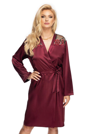 A person wearing an Irall Elodie Dressing Gown in plum, featuring exquisite floral embroidery on the shoulders, stands confidently with one hand on their hip. The luxurious gown by Irall also includes a stylish waist tie, and the individual has long, wavy blonde hair.