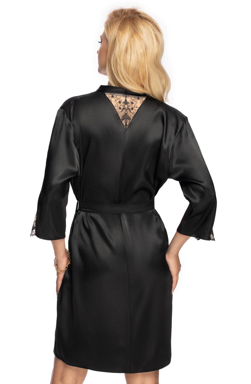 A woman with blonde hair, viewed from behind, is wearing the elegant Irall Mallory Dressing Gown in black. This high-quality satin gown from the brand Irall features v-shaped embroidery and lace detailing on the back and sleeves. She has her right arm relaxed by her side, and the gown is cinched at the waist with a matching belt.