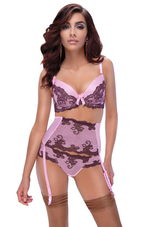 A woman is confidently wearing a coordinated lingerie set, including the Roza Caryca Push Up Bra in pink and brown delicate lace. The set is completed with a matching high-waisted garter belt and thigh-high stockings. She stands with her hands on her hips, looking directly at the camera, her long wavy brunette hair cascading around her shoulders.