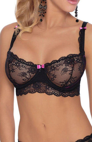 A woman is wearing a delicate Roza Fifi Soft Cup Bra in black, which is adorned with pink bows on the straps and center. The luxurious bra, part of a Roza box set, features intricate floral patterns and scalloped edges. She has long curly blonde hair and wears large dark earrings, with her midsection also visible.