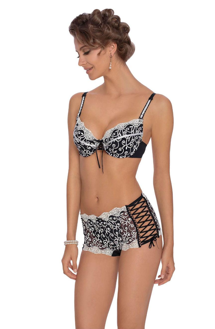 A woman stands smiling, showcasing the Roza Ginewra Short set in black and white with intricate patterns and cream embroidery. The matching push-up bra features delicate lace trim and a small bow in the center, while the high-waisted shorts have elegant lace sides and ribbon detailing. She has curly hair and is accessorized with pearl earrings and a pearl bracelet.