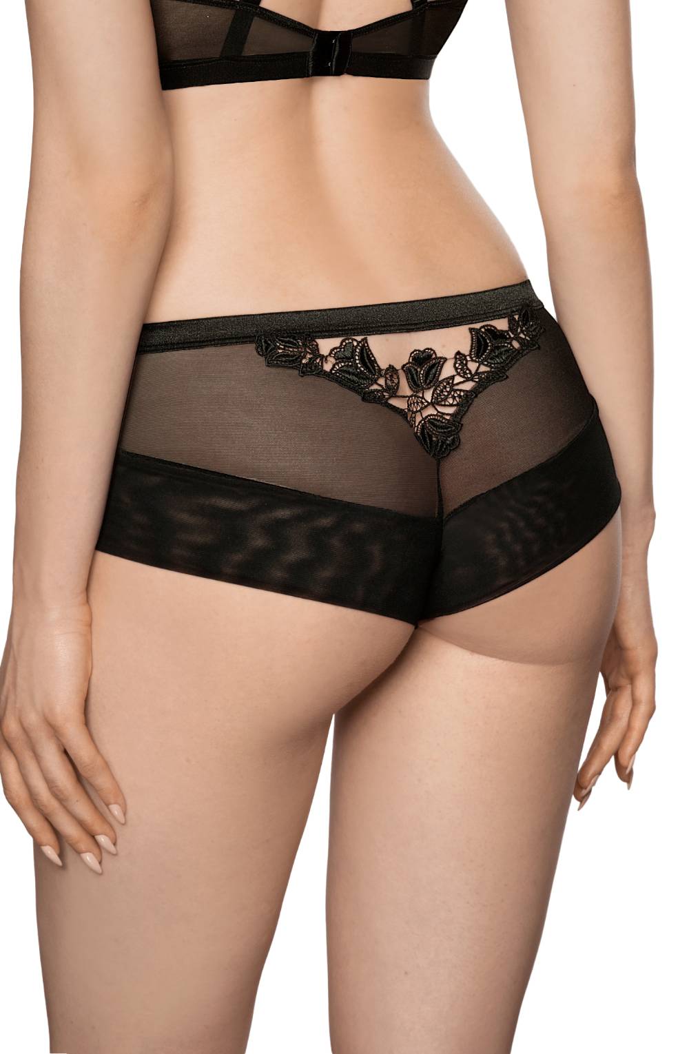 A rear view of a person wearing Roza Lisbet Black Brief lingerie by Roza. The sheer black mesh fabric, adorned with decorative floral embroidery and flower appliques from the collection, gracefully enhances their silhouette. The person's hands rest relaxed by their sides.