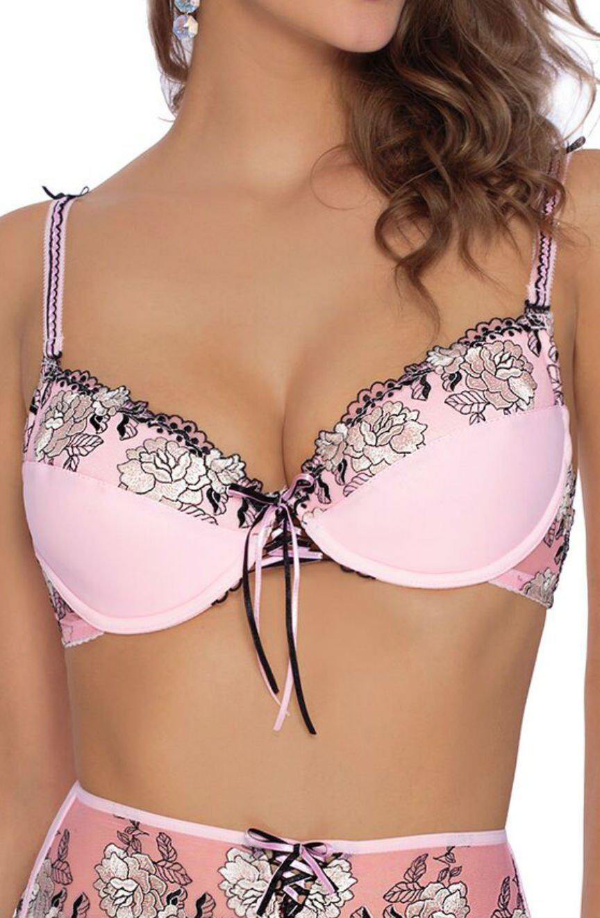 A woman is wearing a Roza Natali Pink Bra by Roza, adorned with black lace trim and floral embroidery. The bra features a black ribbon bow in the center front. She is also wearing matching high-waisted panties with the same decorative pattern. Her hair is styled in loose curls.