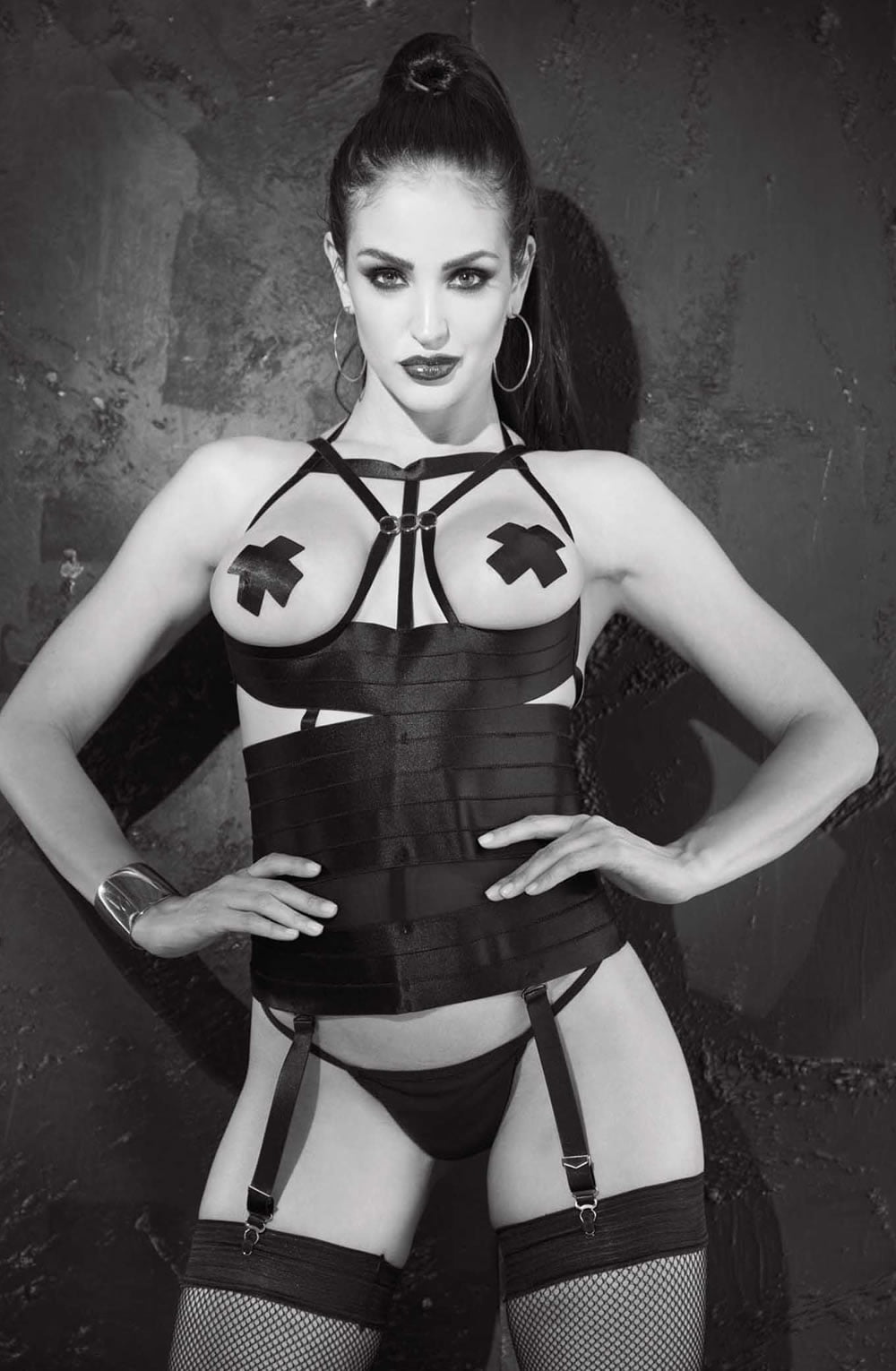 A woman confidently poses against a textured dark wall, wearing the Shirley Of Hollywood 26944 Open Bust Bustier by Shirley of Hollywood. Complementing her look are cross pasties, fishnet stockings, and hoop earrings. Her pulled-back hair and dark lipstick further exude a bold and edgy style in the monochromatic black-and-white image.