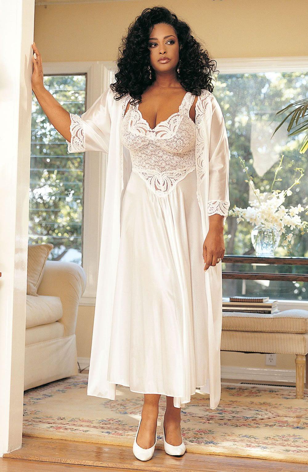 A woman stands in a well-lit room wearing the Shirley of Hollywood X3585 Bridal Gown in white, complete with lace-trimmed detailing and an elegant peignoir set. She has curly hair and is posed with one hand resting on a door frame. The room features a cozy armchair and large windows showcasing a lush green outdoor view.