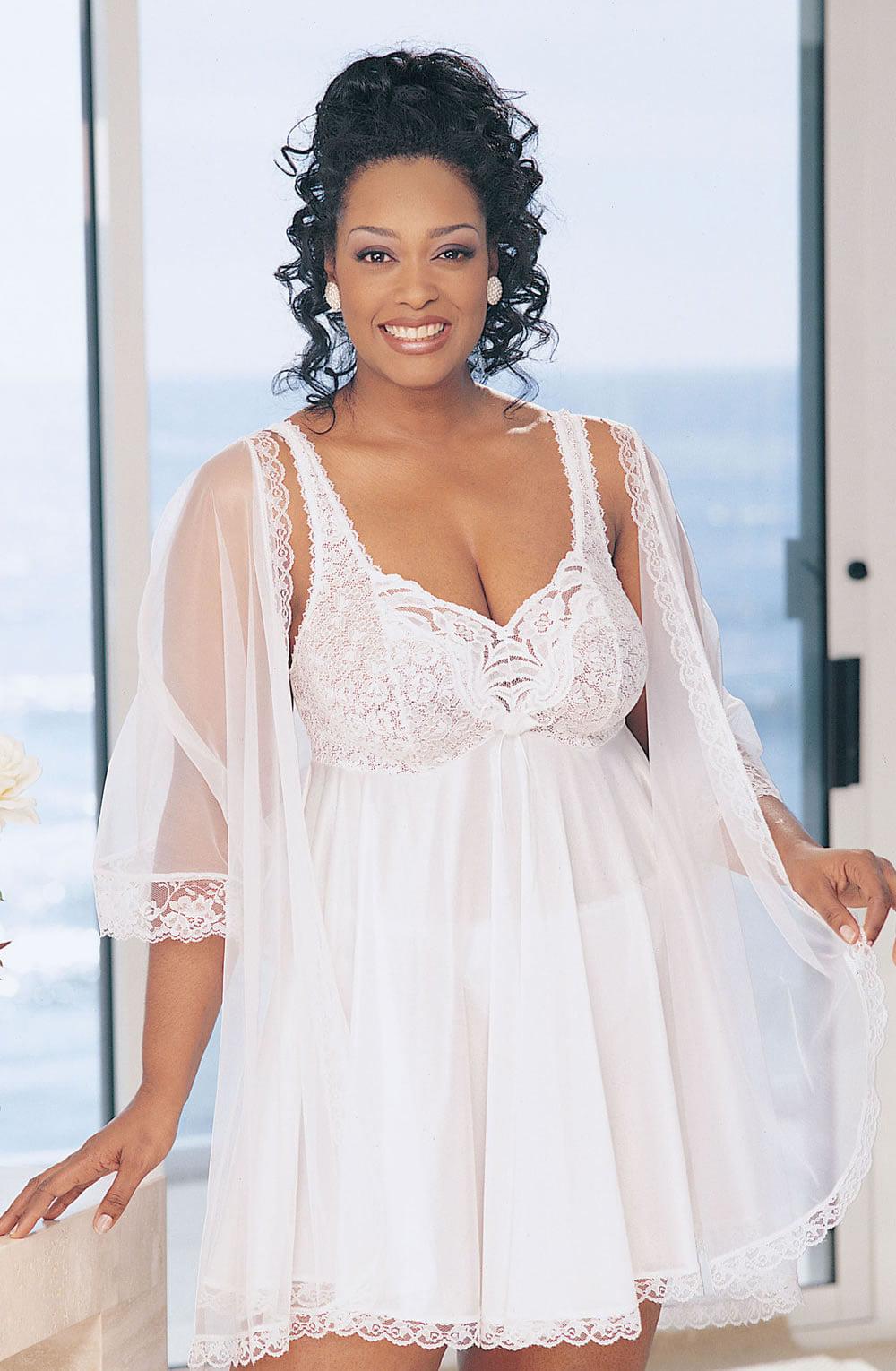 A woman stands by a window with the ocean in the background. She is wearing the Shirley Of Hollywood Xx3595 Babydoll White, a white lace peignoir set with sheer sleeves, adjustable straps, and a flowing skirt. Her curly black hair is styled up, and she is smiling at the camera.