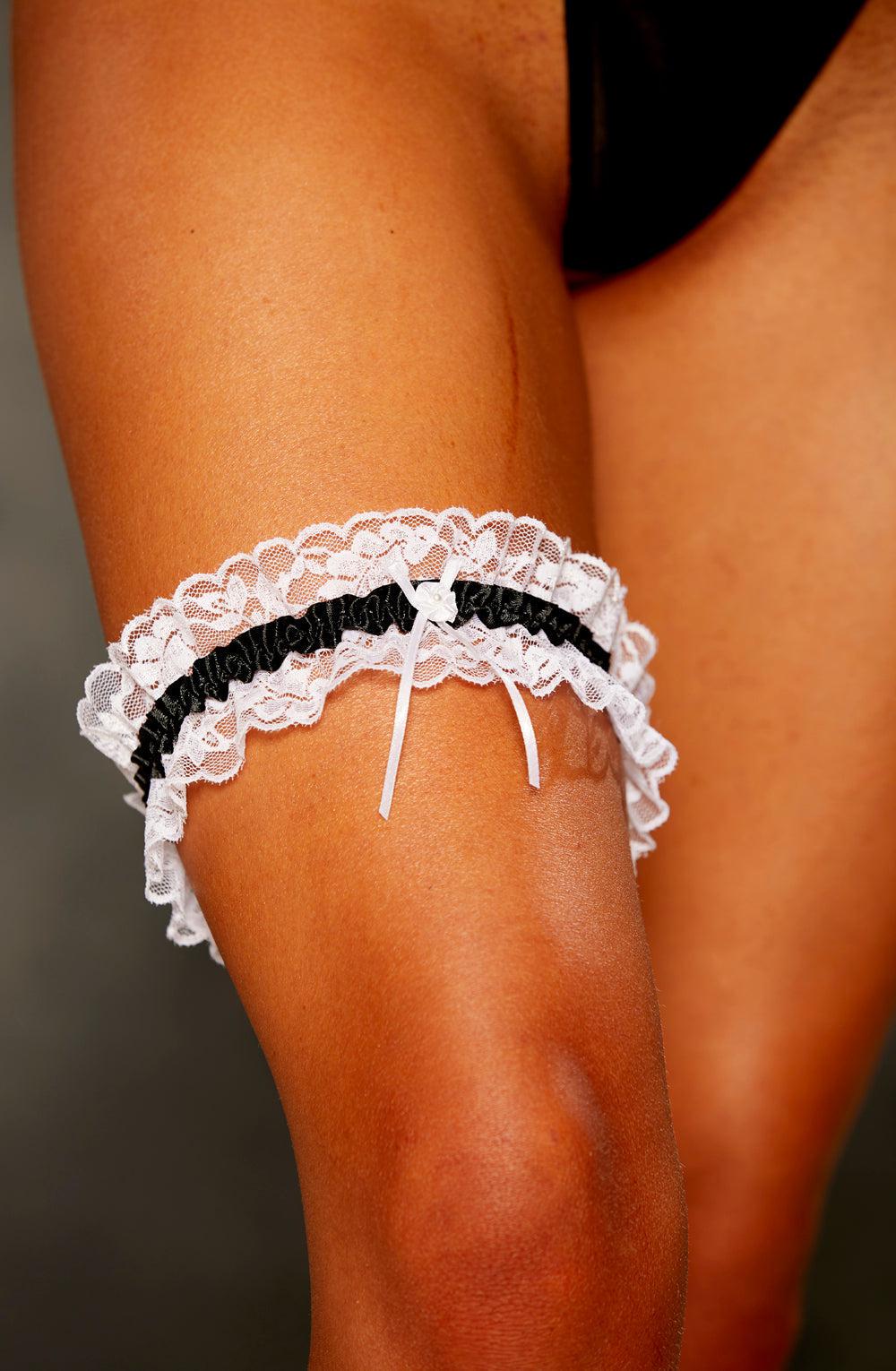 Close-up of a person's thigh wearing the Shirley Of Hollywood 19 Black Garter, which features white lace with black accents and a small white bow. The background is plain, ensuring the garter stands out. The person's skin tone is visible, and they are partially wearing a black garment.