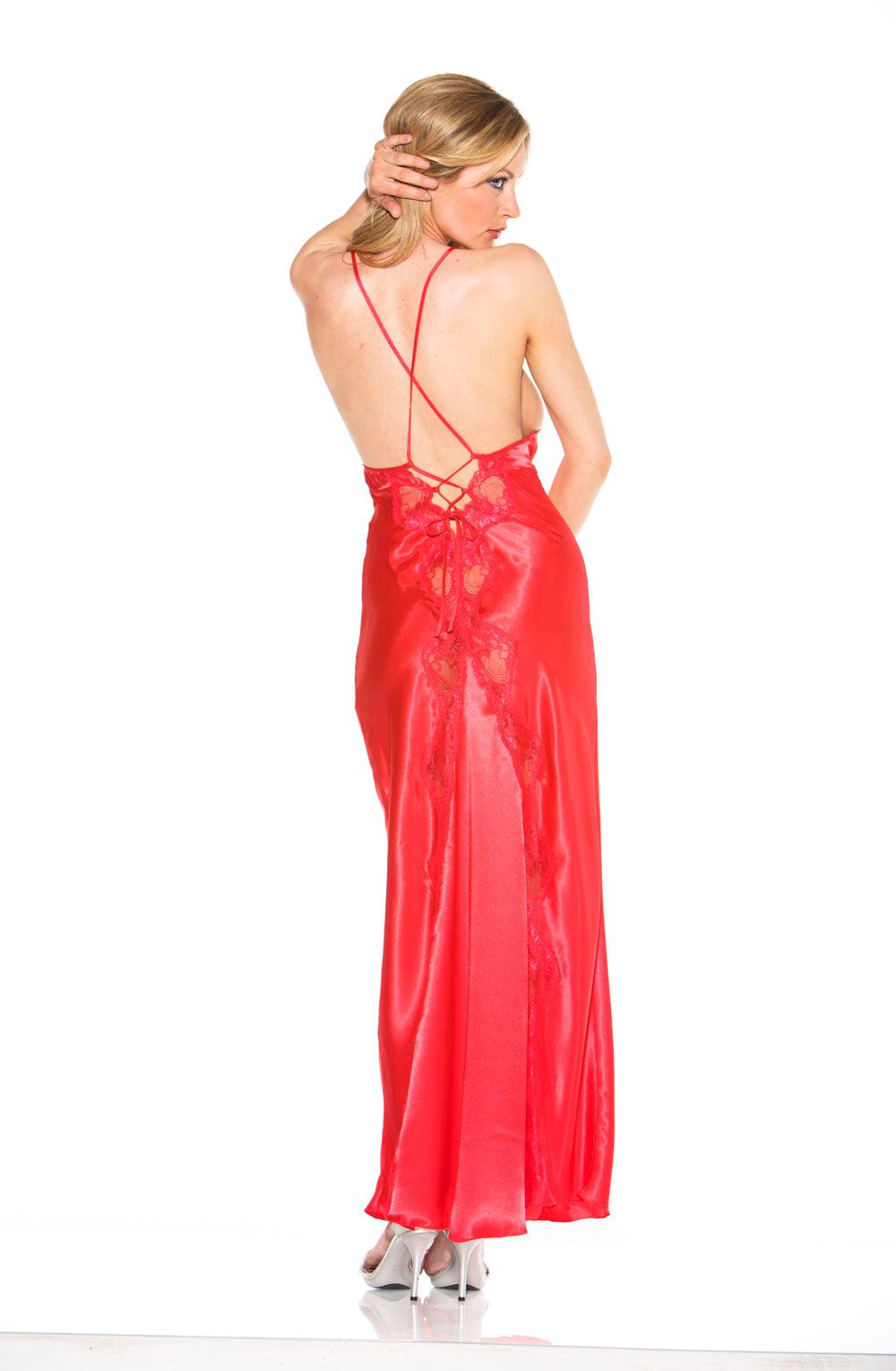 A woman stands posing with her back to the camera, wearing the Shirley Of Hollywood 20300 Red Long Gown by Shirley of Hollywood, featuring a low-cut back and crisscross tie shoulder straps. She is holding her hair with one hand while her right arm rests on her hip. She completes the look with white heeled shoes.